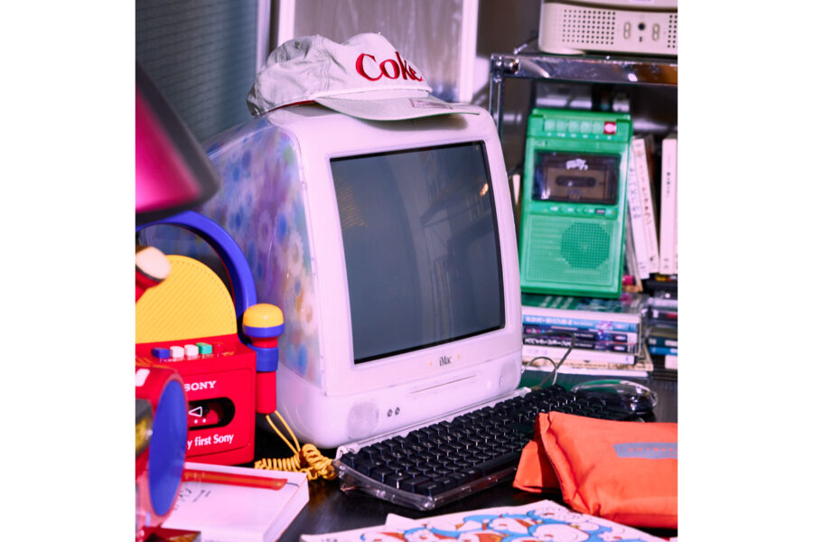 Cluttered retro computer setup with an iMac, keyboard, and colorful toys on a dark desk, cap reading 'Coke' on top of the computer