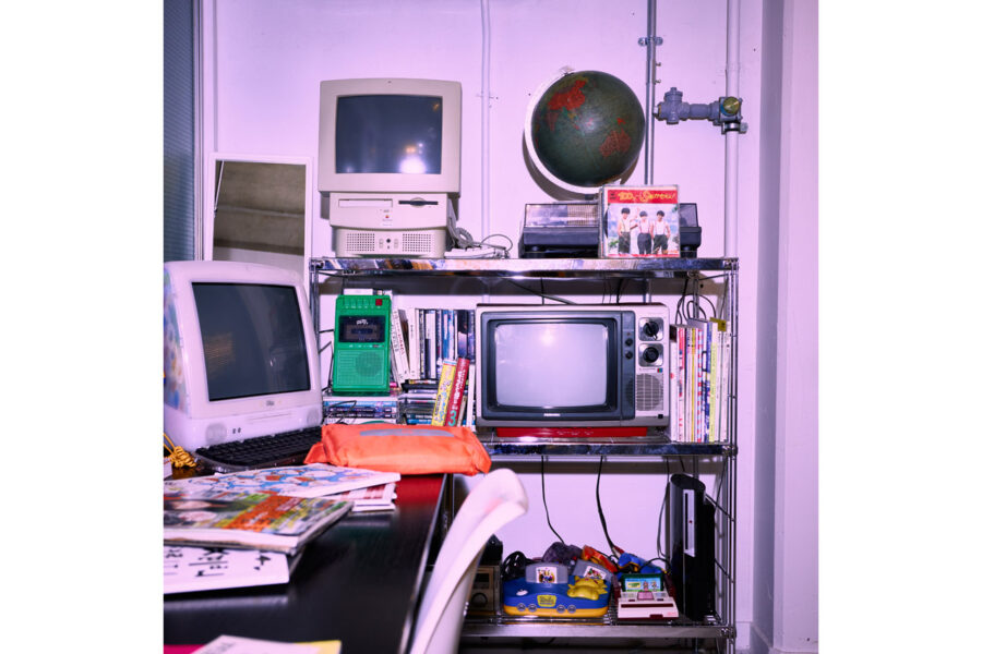 Cluttered retro tech shelf featuring a beige CRT computer, a small TV, a globe, and a stack of books and magazines.