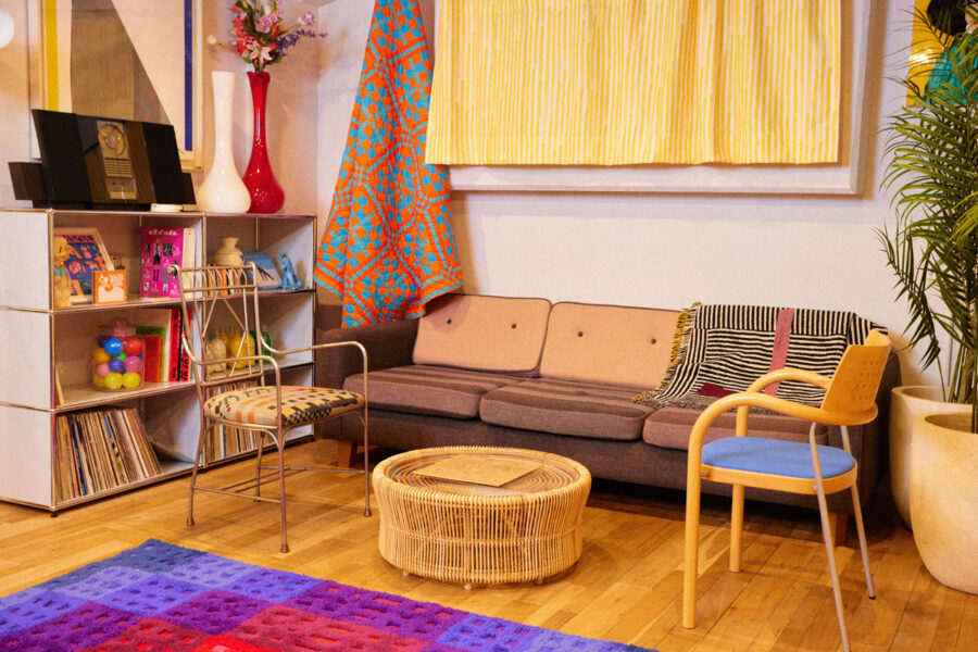 Cozy retro living room with a brown sofa, round wicker coffee table, and a colorful rug amid a bookshelf and plants (kid-friendly setup).