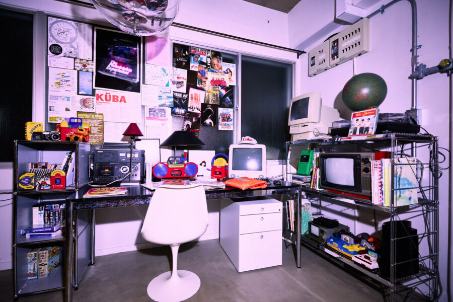 Retro tech workspace with vintage computer, CRT monitor, boombox, and shelves of gadgets against a poster-filled wall.