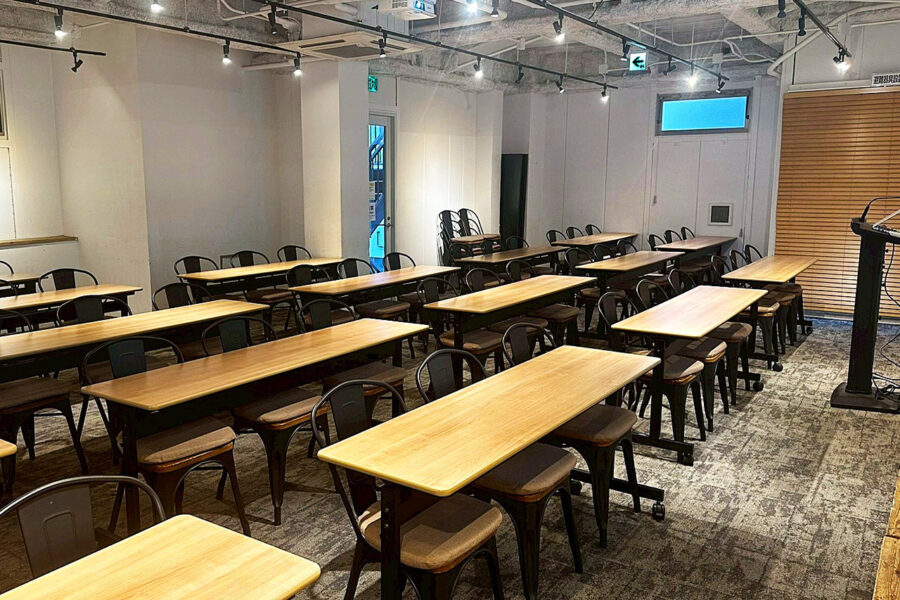 Empty classroom with rows of wooden-topped tables and black metal chairs, and a podium at the front right.