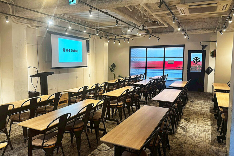 Training room set up for a presentation with long wooden tables, black chairs, a projector screen showing 'TIME SHARING', and a podium under string lights in a modern coworking space.