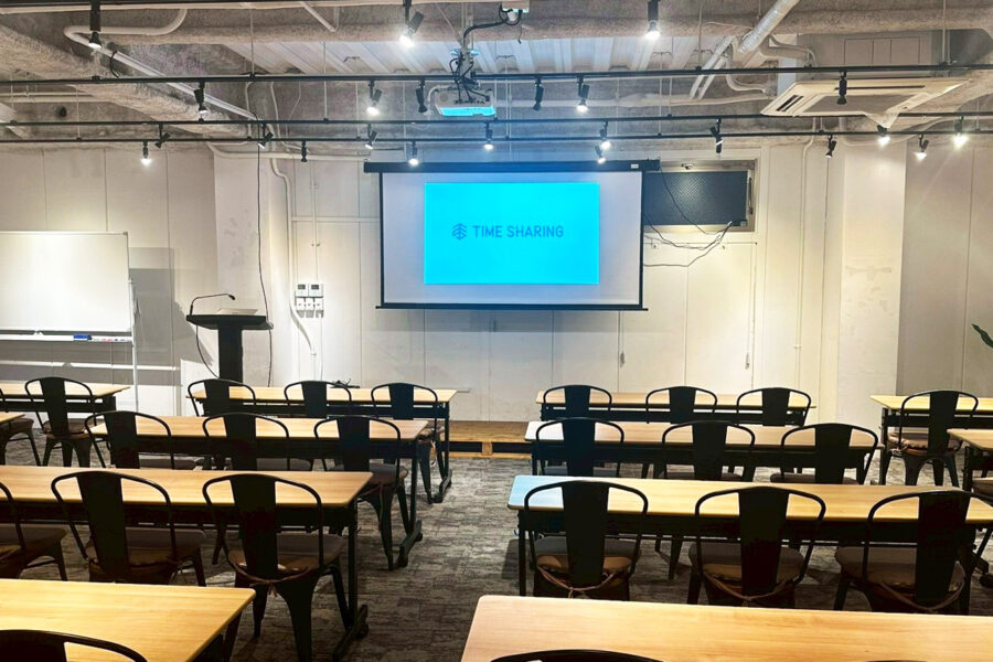 Lecture room with projector screen showing 'TIME SHARING', rows of wooden tables and black chairs, podium at front.