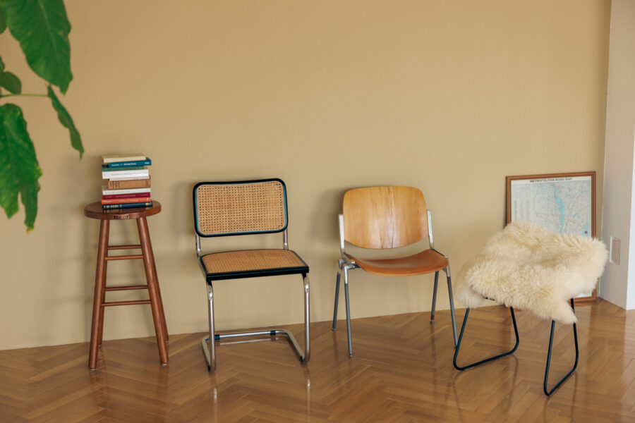 Row of three different chairs along a beige wall, with a fur-covered chair on the right and a framed map resting on the floor to the right. A small stool with books sits on the left, and a green plant is visible in the upper left corner.