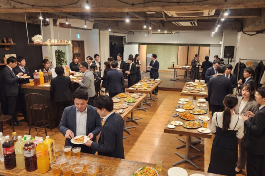 Office reception with dozens of professionals in suits around long buffet tables of food and drinks.