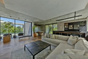 Open-concept living room with a gray sectional, black coffee table, and floor-to-ceiling windows overlooking greenery.