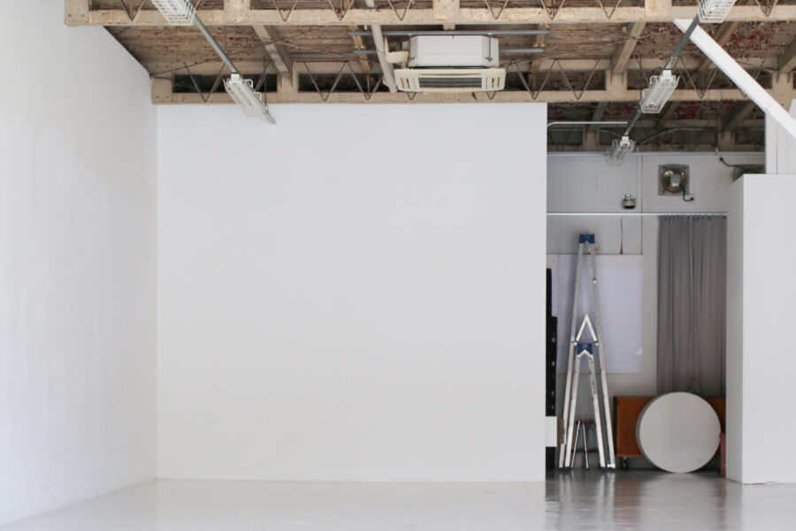 328A5088+++ Empty white photography studio with exposed ceiling beams, a metal ladder in the corner, and a round white tabletop on the floor.