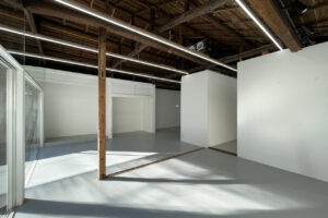 Industrial-style empty gallery space with white walls, exposed wooden ceiling beams, and a glass door on the left.