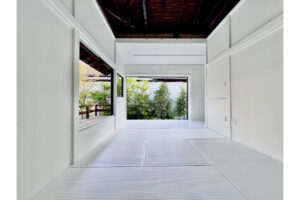 White minimalist interior with tatami-like flooring opening onto a small garden with green trees.