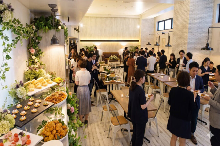 Reception with a long buffet and guests mingling in a bright, stylish venue; floral decor along the food wall and standing tables filled with food and drinks