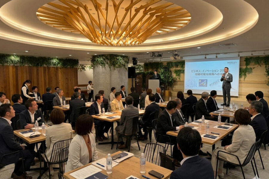 Speaker presenting at a business conference with attendees seated at round tables and a projection screen behind him.