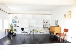 Bright minimalist living space with white built-in shelves, a round table, and mixed chairs on a dark wood floor.