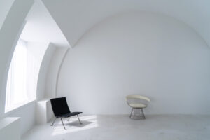 Minimal white room with a curved wall, a large window letting in light, a black lounge chair, and a white wire chair nearby.