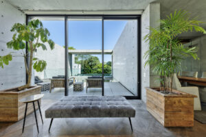 Living room with concrete walls and large glass doors opening to a patio; two wooden planters with leafy trees flank a gray tufted ottoman.