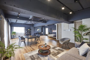Open-concept loft with blue walls and exposed beams, featuring a round dining table, sofa, rug, and indoor plants, with a bedroom area in the back.