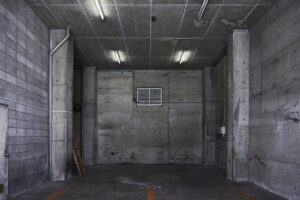 Empty concrete garage bay with exposed pipes, ladder, and fluorescent lights on a damp, industrial wall.