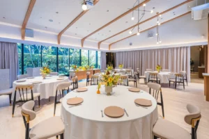 Elegant banquet hall with round tables in white linen, beige plates, yellow flower centerpieces, and large windows.