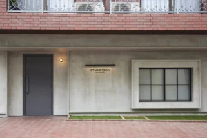 Facade of a modern studio building: gray door left, large framed window right, sign says 'STUDIO PEAK' with smaller text beneath, brick balcony above.