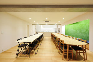 Long communal dining/meeting room with two parallel tables and many chairs, white walls, and a bright green wall at right; kitchen in back.