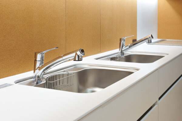 Double stainless steel kitchen sinks with chrome faucets, set into a white countertop with a light brown cork backsplash.