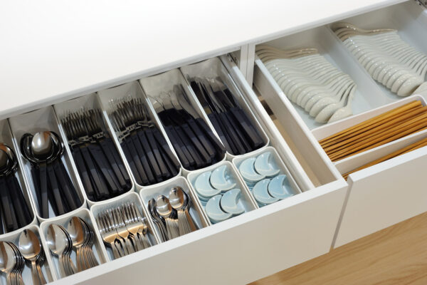 Organized kitchen drawer with separate compartments holding forks, knives, and spoons in black handles, plus light-blue ceramic spoons. Right side shows stacked white plates and chopsticks in a tray.