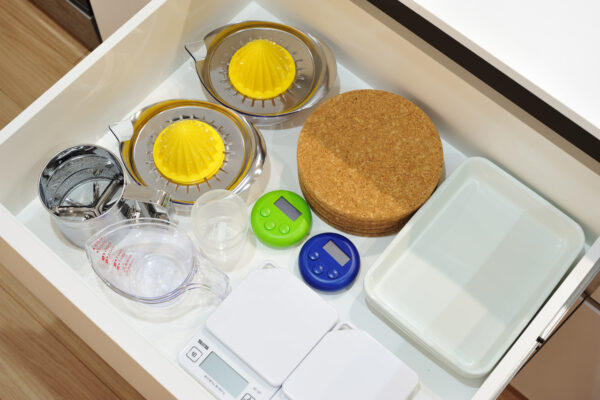 Drawer of kitchen gadgets: two yellow citrus reamers, metal juicer, cork round cutting board, green and blue digital timers, measuring pitcher, white trays, and a scale.