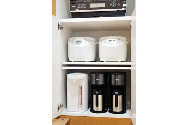 White kitchen cabinet with two white rice cookers on the top shelf and a white kettle and two black coffee makers on the bottom shelf