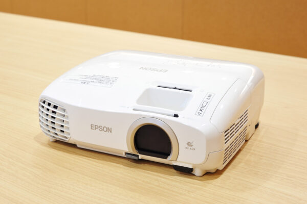 White Epson projector resting on a light wood table, front lens covered by a circular cap, side vents visible.
