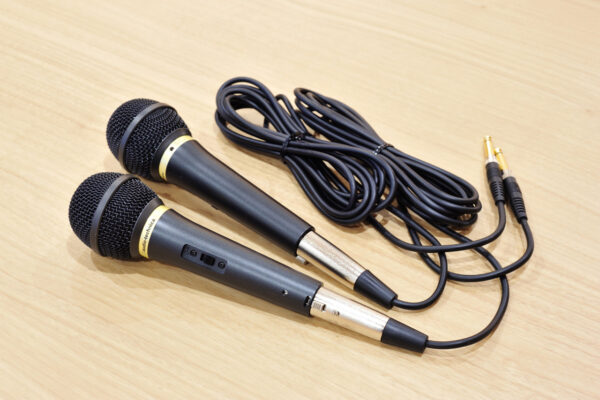 Two handheld wired microphones with black bodies and gold bands lie on a wooden surface, connected to a long black cable with 1/4-inch plugs.