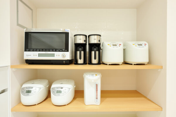 Two-tier pantry-style setup with multiple small appliances: a microwave, two coffee makers, two white rice cookers, and a water boiler with a thermometer on wooden shelves.