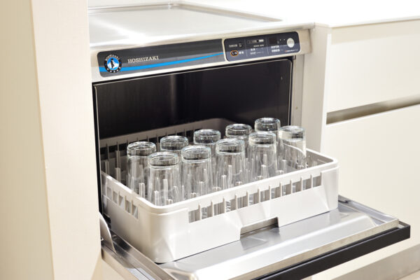 Open dishwasher with a white rack holding tall clear glasses in a kitchen. Brand Hoshizaki is visible on the unit.