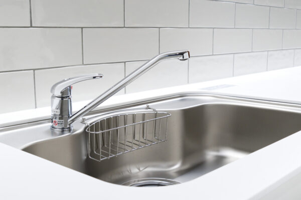 106 Stainless steel kitchen sink with a curved chrome faucet and a wire rack basket in the basin, with a tiled backsplash behind.