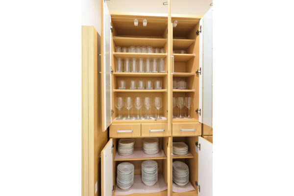 Wood kitchen cabinet with open doors showing stacked glassware on upper shelves and plates on lower shelves.