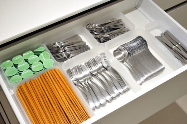 A neatly organized kitchen drawer with compartments holding orange chopsticks, stacked forks, spoons, and knives, plus green round lids in a separate tray.
