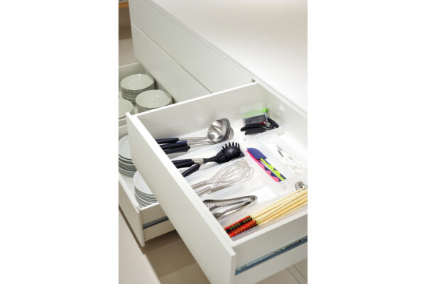 Open kitchen drawer filled with utensils: ladles, whisks, tongs, and chopsticks in a white cabinet.