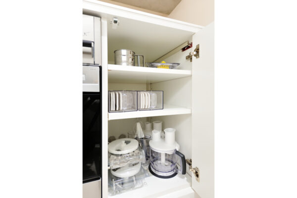 Open kitchen cabinet showing organized shelves: a metal steamer on the top shelf, dishware in a clear box on the middle shelf, and a food processor with attachments on the bottom shelf.
