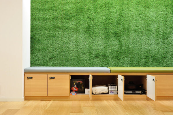 Bench with gray and lime cushions against a green plant wall, wooden storage cabinets with open doors showing electronics inside (printer, control unit, etc.).