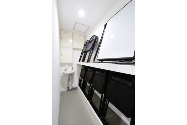 114 Narrow room with stacked black folding chairs along a whiteboard wall and a small sink with cabinets nearby.