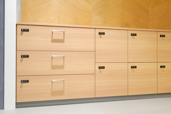 115 Row of light wood cabinets with metal handles and small lock safes on each drawer along a wall with a white baseboard.