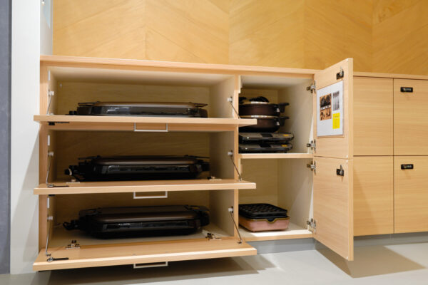 116 Open wooden kitchen cabinet with four pull-out shelves holding several electric grills; right-side door shows stacked pans and a pink grill pan.