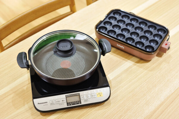 119 Induction cooker with a pot and glass lid on it, and a takoyaki pan nearby on a wooden table.