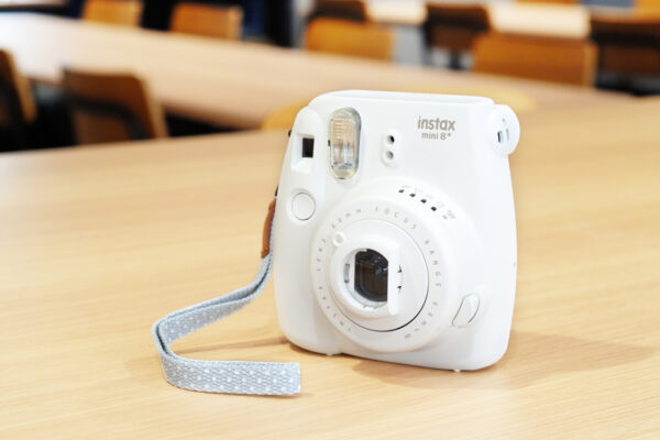 120 White Instax mini 8+ instant camera on a light wooden table with a pale blue patterned strap resting nearby.