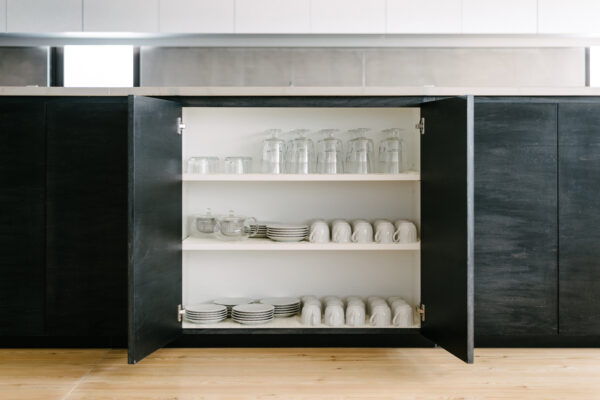 Open kitchen cabinet with glassware, cups, and plates neatly arranged on white shelves in a dark cabinet setting, wooden floor