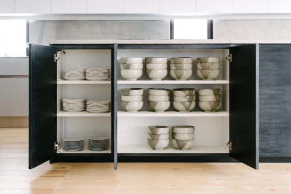 Open kitchen cabinet with stacked white plates on the left and beige ceramic bowls on the shelves in the center and right.