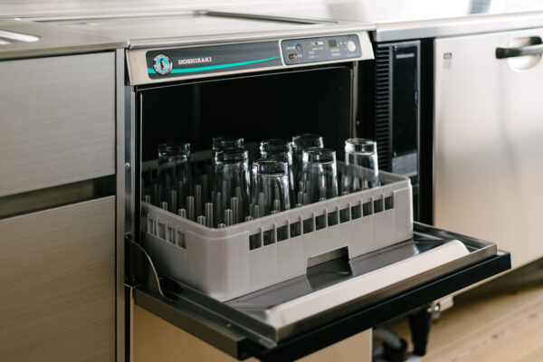 Open dishwasher with a gray plastic rack holding tall drinking glasses, ready for washing in a stainless steel kitchen.
