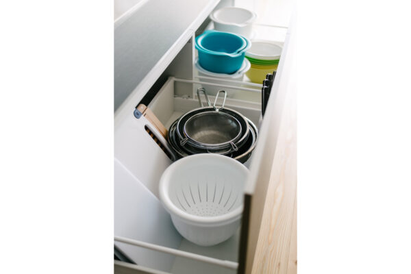 Open kitchen drawer containing metal strainers, a white colander, and colorful bowls stacked in the back.