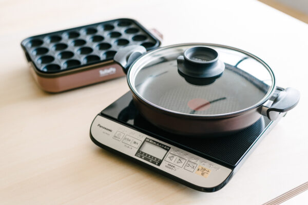 Electric induction cooktop with a glass-lidded pot; a takoyaki pan sits nearby on a light wooden table (Panasonic controls visible).