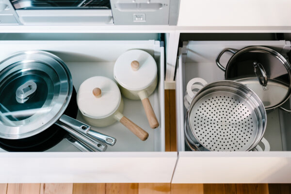 Open kitchen drawer showing white pots with wooden lids and handles alongside stacked stainless-steel pans and lids in a built-in drawer.