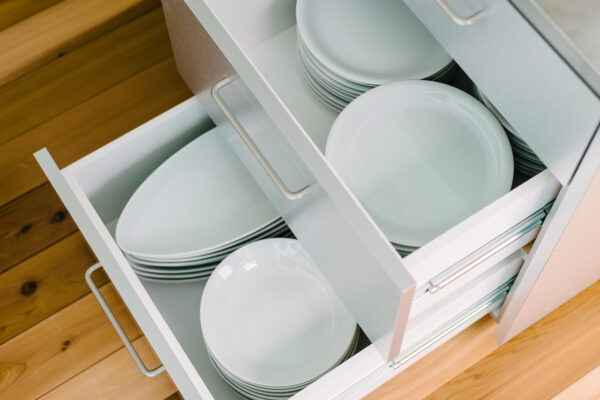 Open kitchen drawer showing stacked white plates and bowls inside a light cabinet on a wooden floor.