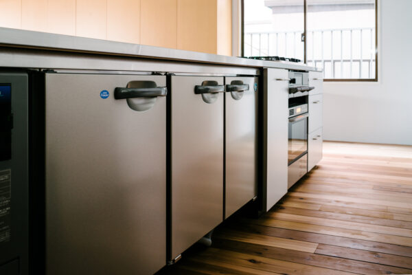 Row of stainless steel kitchen appliances, including under-counter fridges and an oven, along a wooden floor in a modern kitchen
